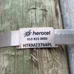 The photo is close up of a weathered wooden pole wrapped with a galvanized metal strapping. Affixed to that small white plastic tag. The tag carries a black sunburst style logo and the word herotel along with a telephone and pole number.